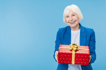 Aged senior woman holding presents gift box red package feels happy looks at camera isolated on blue background. Celebrating New Year Christmas concept