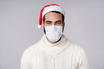 Serious middle-eastern arabian man in protective face mask against Coronavirus Covid19 in Christmas New Year red hat looking at camera isolated over grey background