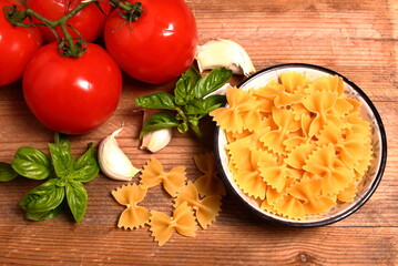 Farfalle raw Italian pasta in a bowl on wooden background with tomatoes garlic and basil. View from above.
