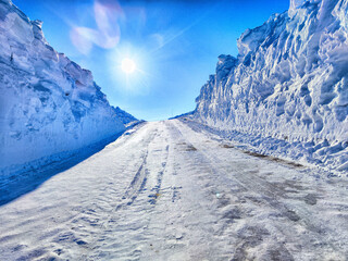 A snow-covered road with towering snowbanks under bright sunshine in a winter landscape, offering serene beauty and solitude