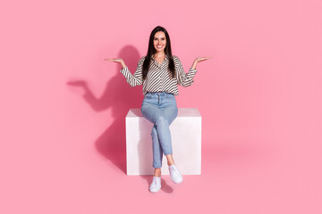 Cheerful young woman in casual style sitting with open arms on a pink background, promoting a...
