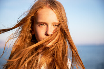 Portrait of a beautiful teenage girl with long flowing red hair against the background of the sea in summer at sunset.