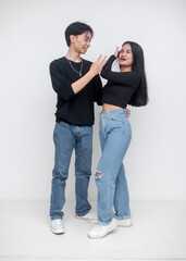 A young asian man and woman in matching black shirts and jeans having a lighthearted conversation, against a white background. Whole body image.