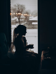 Silhouette of a beautiful bride with cap of coffee in a traditional white wedding dress near window.