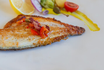 Grilled sea bass in a white porcelain plate. Garnish: lemon, onion, tomato, capers, broccoli, potatoes, carrots.