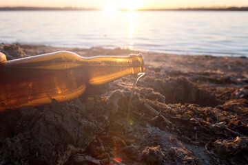 Glass bottle littering a sandy lakeshore at sunset, symbolizing environmental pollution and the...