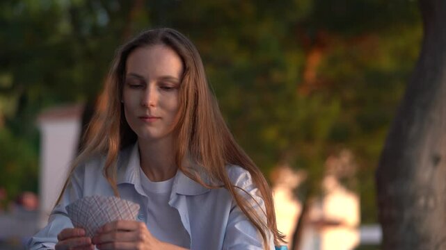 A young woman holds cards and chooses how to suit, looks at trump card, and puts card on the deck at the table outdoors. The concept of board card games, having fun, entertainment, poker, solitaire