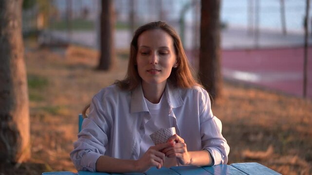 A young woman holds cards and chooses how to suit, looks at trump card, and puts card on the deck at the table outdoors. The concept of board card games, having fun, entertainment, poker, solitaire