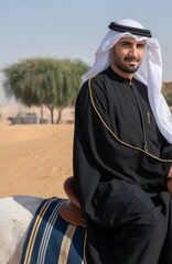 A photo of an Arab Sheikh wearing a black abaya and a white head covering. He is sitting on the back of a white horse. The background contains a sandy desert landscape with a few trees and buildings
