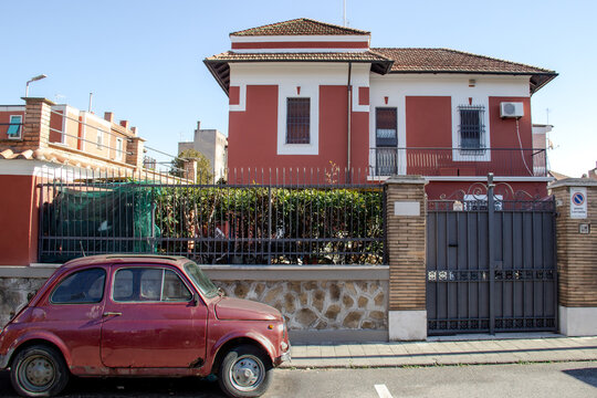 Old rusty red car in front of a beautiful new house. Сontraposition. Pigneto, Rome, Italy, 2023. 