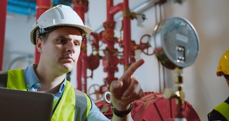 Engineers Analyzing Equipment inspects industrial piping while using a laptop in a Factory, Focused...