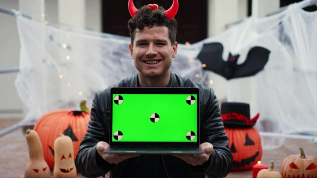 Young man dressed as a vampire devil with makeup holds a laptop with a green screen with dots for tracking movement and pumpkin on the stairs of the porch of the house against the background of
