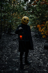 The man Pumpkinhead Jack, stands in the woods in a cloak with a lantern and a red candle.