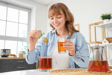Young woman adding sweet honey into cup of tea in kitchen