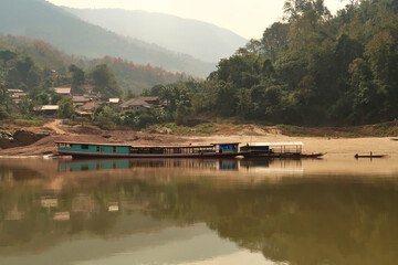 View from the slow boat from Thailand to Laos onto the shore of the Mekong River, another docked slow boat is reflecting in the smooth water in front of a local village, Laos