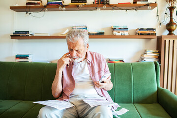 Elderly man reviewing bills and using smartphone on the couch