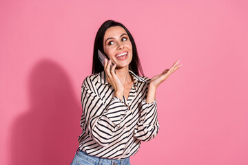 Joyful young woman with brunette hair in casual striped shirt talking on phone against pink background