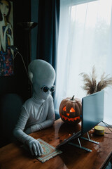 Teenager in Halloween full-body freaky white costume with carved pumpkin laptop on table studying online