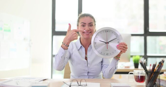 Businesswoman holds large wall clock pointing with finger sitting at desk. Smiling lady hints about time management skills for successful career