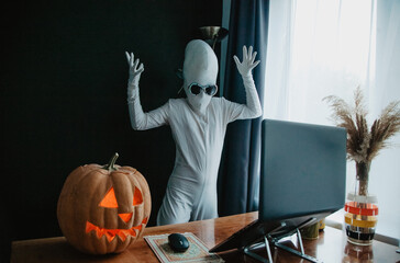 Teenager in Halloween full-body freaky white costume with carved pumpkin laptop on table studying online