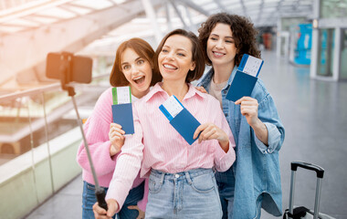 Three enthusiastic friends stand together at the airport, holding their tickets, smiling, and...