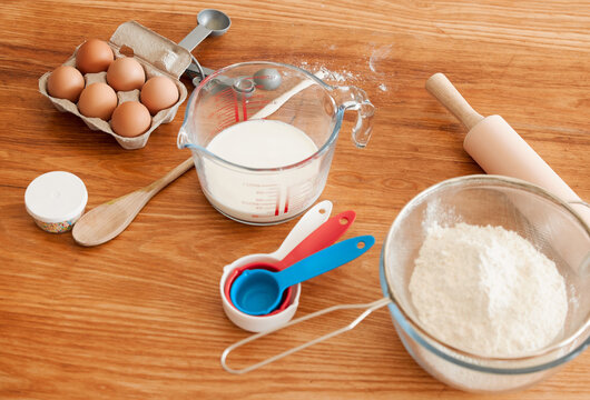Utensils, baking ingredients and eggs on kitchen counter with flour, container and spoon in home. Bowl, milk and rolling pin for process, cooking and prepare for dessert, cookie or cake at apartment