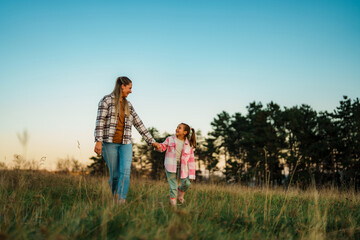 Fototapeta premium A mother and daughter walking hand in hand through a grassy field at sunset, with trees in the background.
