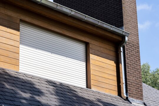 A close-up of a house exterior showing a white roller shutter window framed by wood panels and a brick chimney. The gutter system runs along the edge of the roof, casting shadows under the sunlight.