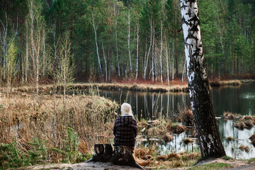 In the image, a man is sitting with his back to the viewer by the water, surrounded by a forest. Trees, water and vegetation are visible. A peaceful natural scene.