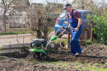 A farmer in the garden tills the land with a motorized cultivator or power tiller, preparing the soil for planting crops