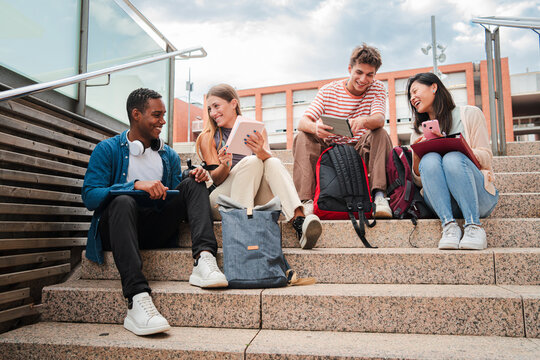 Group of diverse young students talking and smiling while sitting on university campus stairs. Multiracial friends enjoying casual conversation, holding tablets and smartphones in a relaxed academic