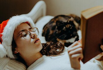 A girl in a Santa hat reads a book with a cat.