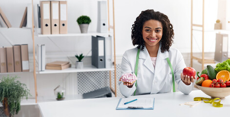 Pretty black woman dietary doctor holding donut and apple, posing at her office, healthy food...