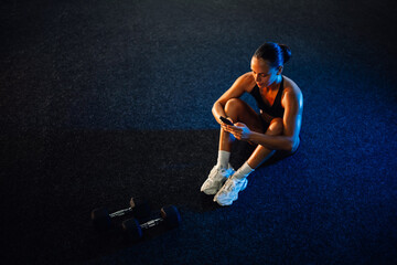 Young athletic woman using smartphone app after training with dumbbells