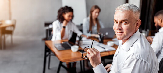 Business Career. Senior Businessman Sitting At Corporate Meeting With Employees Smiling To Camera In Modern Office. Copy Space