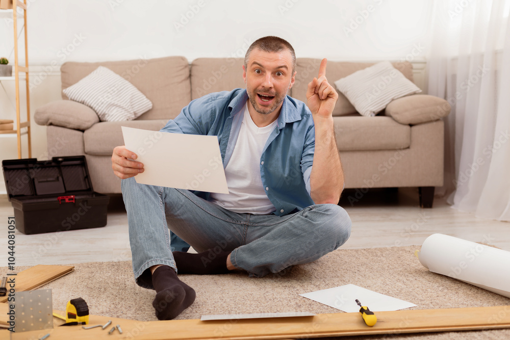 Poster excited man assembling furniture pointing finger up having idea sitting on floor at home, reading in