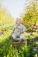 A cute baby boy is sitting on the grass in the park and smiling. A stylish baby in a beige romper.