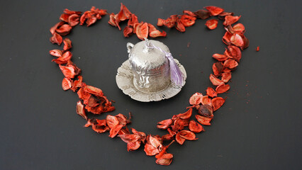 Silver coffee cup set placed at the center of a heart shape made with red dried flower petals on a...