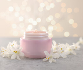 Cream in jar and beautiful jasmine flowers on grey table against beige background with blurred lights, closeup