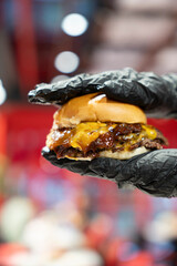 A person wearing black gloves is holding a burger with melted cheese and sauce.