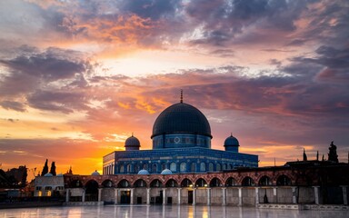 Obraz premium A blue domed mosque with intricate tile work stands against a backdrop of a vibrant sunset sky.