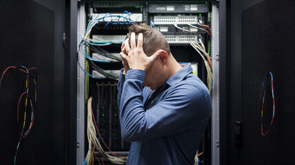 A man in a grey shirt stands in a server room, looking concerned and holding his hand to his forehead. Behind him are several server racks with various cables and connections.