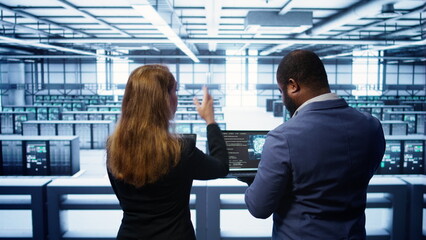 Teamworking programmers in data center overseeing server operations using laptop and tablet. Coworkers ensuring optimal performance and data integrity in server room using devices, camera B