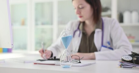 Brunette woman doctor sits at desk in medical office writing report. Hourglass with sand on table decorates workspace in consultation room