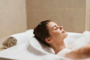 Relaxed young woman enjoying a bubble bath in a modern bathroom, surrounded by soft towels and...