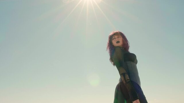 Low angle shot of a young Goth woman, with pale sky and sun flare in the background. Gothic girl strides through the barren landscape with her head turned back, as if looking at something left behind.