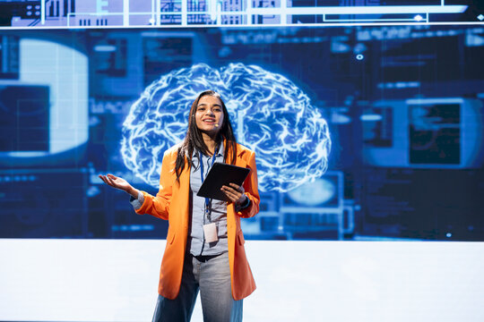 Jolly presenter talking at AI expo using mic and tablet, sharing industry expertise to audience. Upbeat programmer holding device, exploring latest AI products and services at tech event