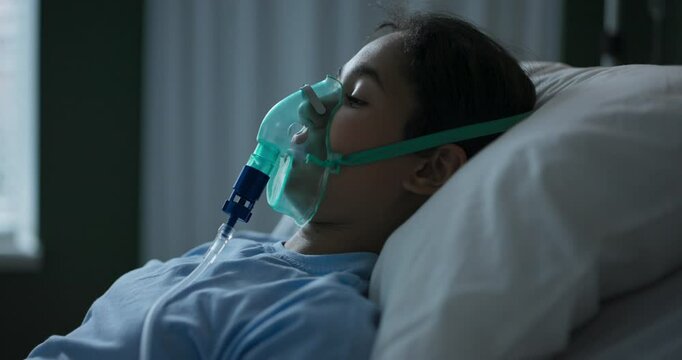A young African girl lies in a hospital bed wearing an oxygen mask after a major surgery.