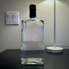 Square sided, clear glass bottle of alcoholic spirt with plain white label, on dark table with light switches on wall behind