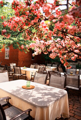 Beautiful outdoor seating area with tables and chairs on a brick patio. Each table, covered with a white tablecloth, has a small green glass jar on a wooden tray.